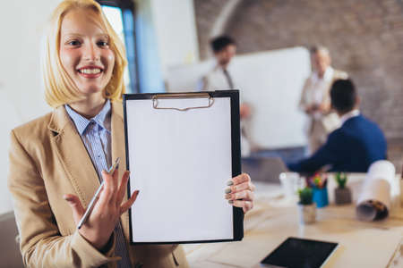 Happy young businesswoman holding clipboard with team discussing project in the background.