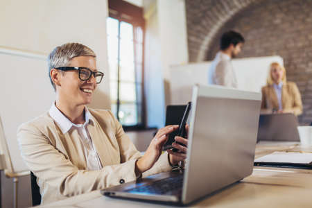 Happy businesswoman using laptop and phone with team discussing project in the background.の写真素材