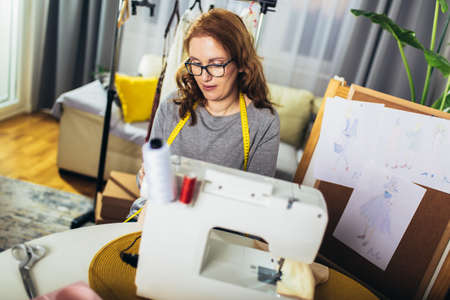 Cheerful young woman sewing while sitting at her working place at home