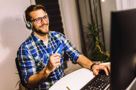 Man sitting at his desk late at night, using computer and credit card for paying online.
