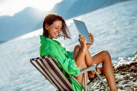 Smiling woman sitting on deck chair by the sea using tablet on a sunny dayの写真素材