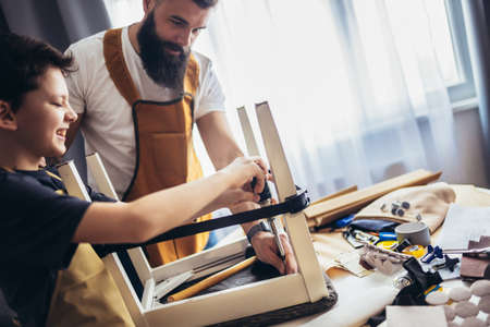 Smiling father is teaching his son upholstery at home, having fun.
