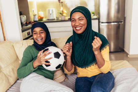 Football match watching concept. Muslim woman and young daughter sit couch cozy celebrate soccer team victory.の写真素材