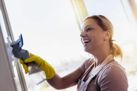 Happy Beautiful Woman wearing Protective Gloves Cleaning Window by spraying Cleaning Products and wiping with Sponge.の写真素材