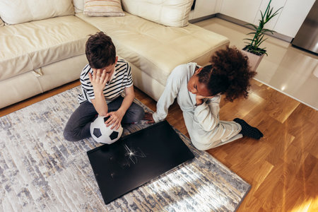 Sad children standing in front of a TV with broken screen holding a ball. Home insurance concept.の写真素材