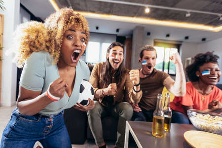 Young group watching sports on television and cheering. Emotional football fans watching football game together at home. Sports Fans with Painted Faces Sitting on a Couch Watch Game on TVの写真素材