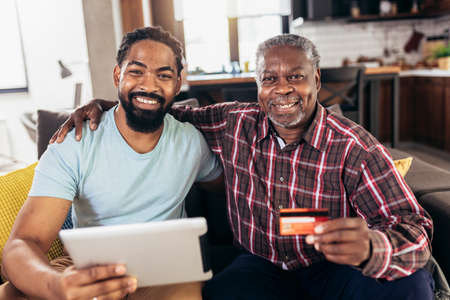 Smiling father and son using digital tablet and credit card for online payment in living room at homeの写真素材