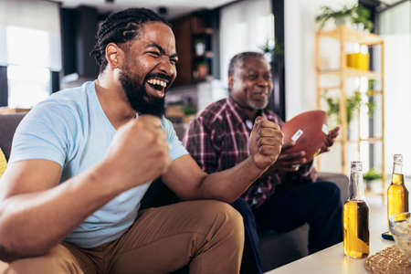 Senior father and adult son cheering for american football game at homeの写真素材