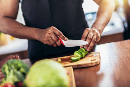 Close up shoot of black man holding sharp knife cutting cucumbers  on wooden board, making fresh salad using organic ingredients.の写真素材