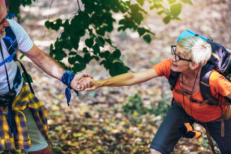 Caring man helping his wife climbing mountain.Senior couple hiking in forest wearing backpacks and...