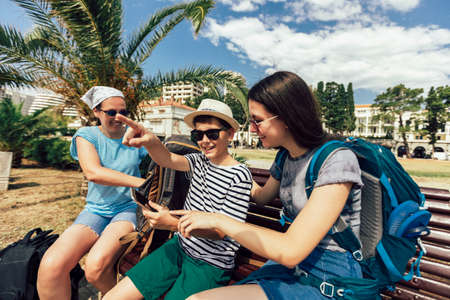 Happy mother, son and daughter together at sea cost with luggage backpack using phone. Travel, tourism, family concept.の写真素材