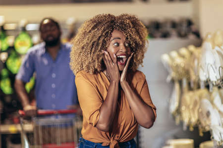 African American couple with trolley purchasing dishes in supermarket. The woman looks excited while...