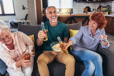 Group of senior friends having popcorn and beer watching tv together during weekend at homeの写真素材