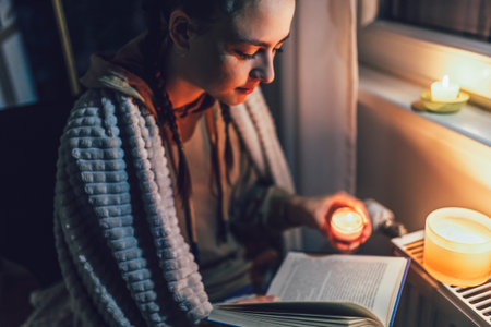 Teenage girl sits under blanket near heating radiator with candles and read book .Rising costs in private households for gas bill due to inflation and war, Energy crisisの写真素材