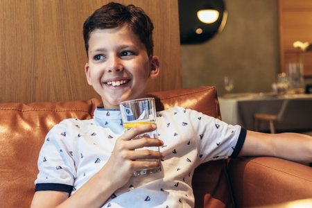 Happy child boy holding glass of fruit juice while waiting for food in restaurantの写真素材