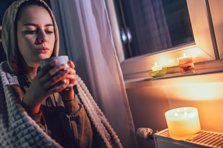 Teenage girl sits under blanket near heating radiator with cup of tea.Rising costs in private households for gas bill due to inflation and war, Energy crisisの写真素材