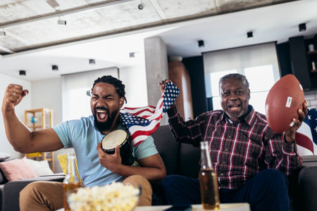 Senior father and adult son cheering for american football game at homeの写真素材