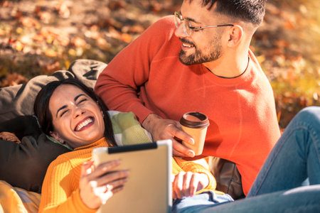 Young man and woman sit on a blanket in the park in autumn using digital tablet.の写真素材