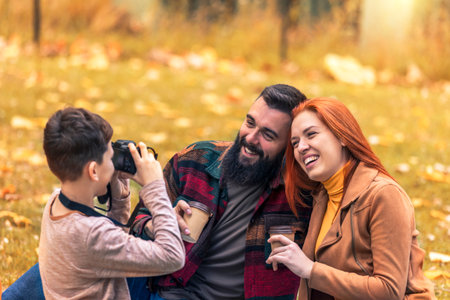 Young happy parents having fun with their boy in the park during autumn day. Boy takes a photo of his parents on a picnicの写真素材