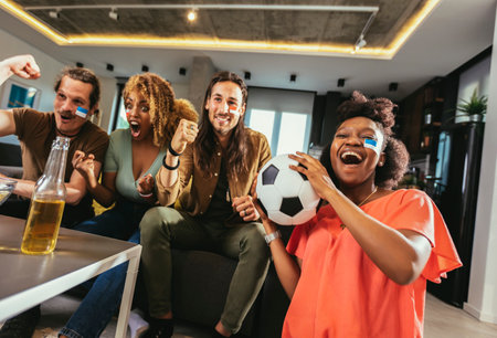 Young group watching sports on television and cheering. Emotional football fans watching football game together at home. Sports Fans with Painted Faces Sitting on a Couch Watch Game on TVの写真素材