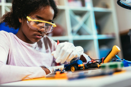 Smart Young African American Schoolgirl is Studying Electronics and Soldering Wires and Circuit Boards in Her Science Hobby Robotics Project. Girl is Working on a Robot in Her Room. Education Concept.の写真素材