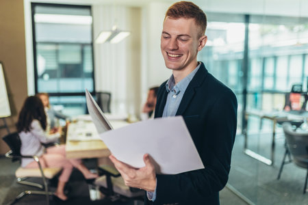Happy businessman standing in the office with team discussing project in the background.の写真素材