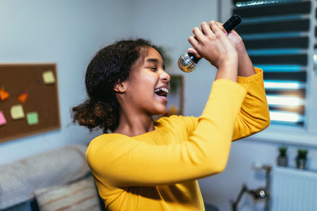 Cute preteen black girl  holding microphon singing karaoke at home, recording songs for contest. Children's lifestyle conceptの写真素材
