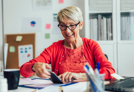 Happy senior woman using mobile phone while working at home with laptopの写真素材