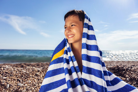 The little boy on the beach caped with a Striped blue and white beach towel and smiling happily.の写真素材
