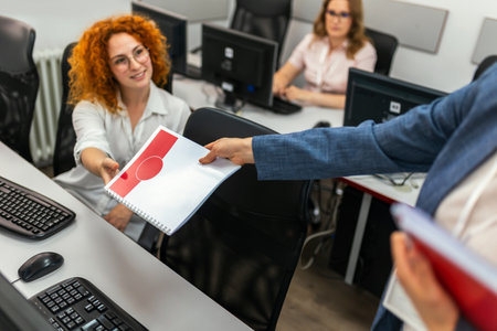 A group of students each sit in front of a desktop in a computer lab. They are each focused on their screens as their female teacher gives them scripts for class.の写真素材