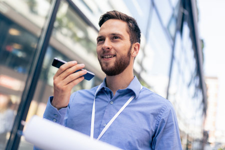 Caucasian bearded male sending voice messages outside of his job. He is using technology for his workの写真素材