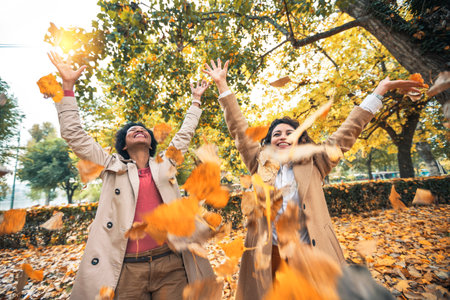 Two smiling young women friends throwing leaves in the park full of fall colors. They are laughing and having funの写真素材