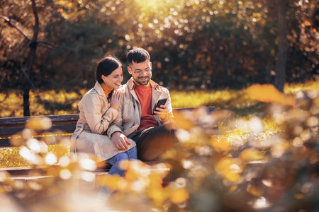 Young couple looking at their tablet. They are sitting on a bench on a sunny autumn day in the colorful parkの写真素材