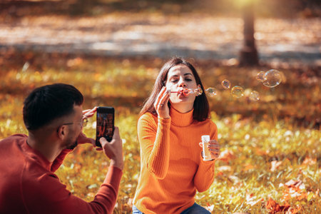 Boyfriend taking a picture of his girlfriend while she blows bubbles. Sunny autumn day in the...