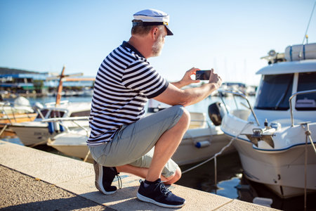 Mature man standing near the sea dressed in a sailor's shirt and hat using smart phone