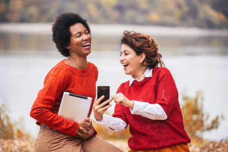 Two happy female friend talking and sharing life stories, enjoying autumn outdoors, using digital tablet and phone. Women's friendshipの写真素材