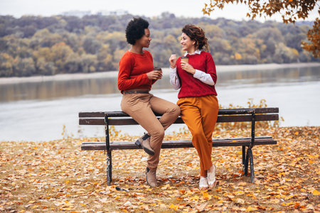 Two happy female friend talking and sharing life stories, enjoying autumn outdoors , drinks coffee. Women's friendshipの写真素材
