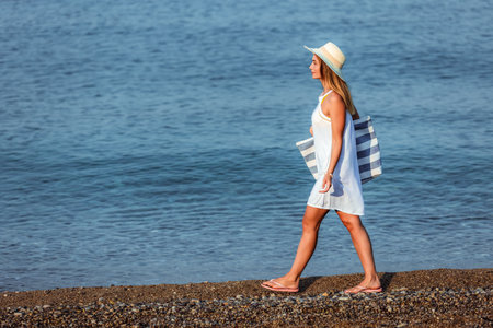 Happy young beautiful woman walking alone on the beach with beach bag and hat.の写真素材