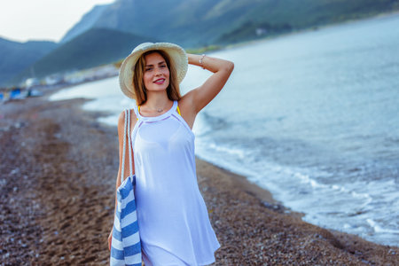 Happy young beautiful woman walking alone on the beach with beach bag and hat.の写真素材