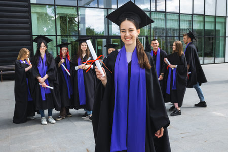 Portrait of graduate student standing in front of her friends with toga and diploma all happy.の写真素材