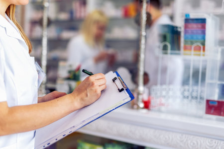 Young female pharmacist standing at the cash desk and writing something on the clipboard in the pharmacy.の写真素材
