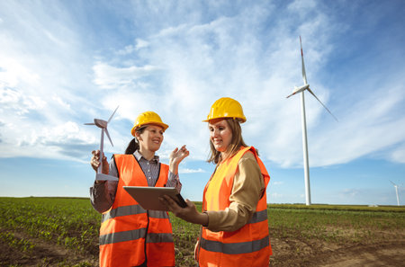 Two women engineers working and looking at the report wind turbine farm. They are wearing florescent vests and helmets for protection.の写真素材
