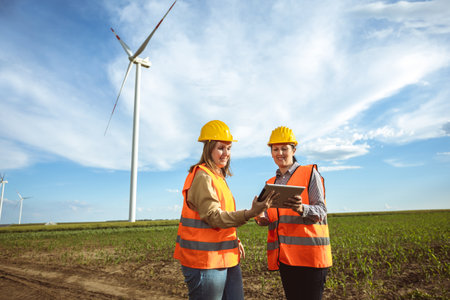 Two women engineers working and looking at the report wind turbine farm. They are wearing florescent vests and helmets for protection.の写真素材
