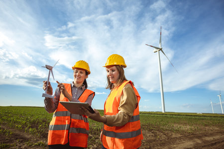 Two women engineers working and looking at the report wind turbine farm. They are wearing florescent vests and helmets for protection.の写真素材