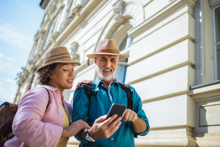 Elderly married couple explores new city. Using their phone for navigation and maps. They are wearing hats and backpacks.の写真素材