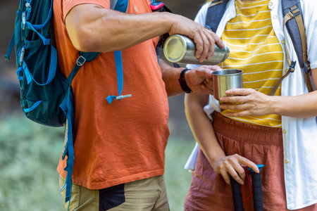 Married couple having coffee break from hiking. They are pouring coffee from a flask and they are also carrying backpacks and hiking poles.の写真素材