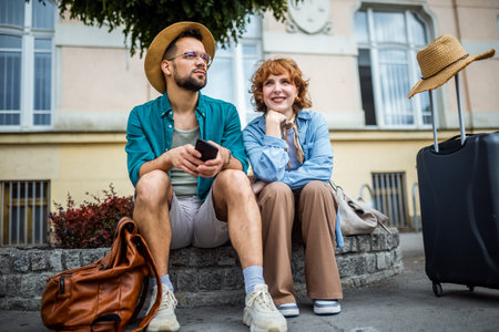 Happy couple sitting in the city center. They are using their phone so they don't get lost. Having their luggages and backpacks with them.の写真素材
