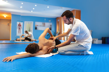 The boy is doing kinesiotherapy exercises with the chiropractor helping him. Exercises for correcting the spine and pelvis, scoliosis. He is lying on his side.の写真素材