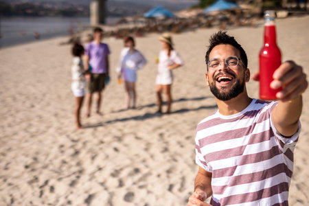 Young handsome man drinking red drink at the beach while his friends are hanging out behind him.