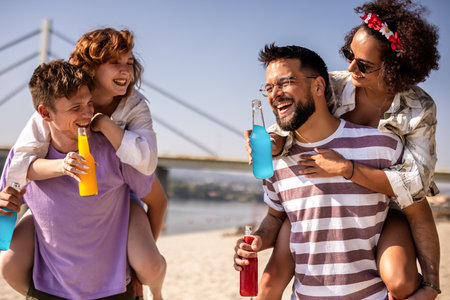 Two male friends are carrying their girl friends on their back on a beach. They are fooling around and drinking cocktails.の写真素材